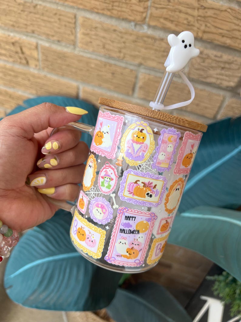 May include: A clear glass mug with a bamboo lid and a white ghost-shaped straw topper. The mug is decorated with Halloween-themed illustrations in frames, including pumpkins and ghosts. The words "Happy Halloween" are visible on the mug.