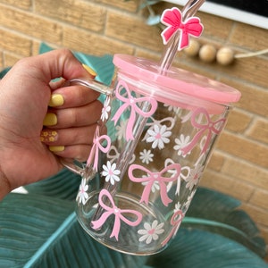 May include: A pink glass mug with a clear straw and a pink bow on top. The mug has a floral design with pink bows and white flowers.
