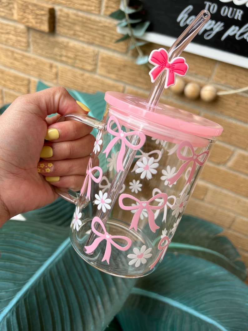 May include: A pink glass mug with a clear straw and a pink bow on top. The mug has a floral design with pink bows and white flowers.