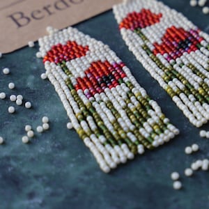 Könnte beinhalten: Ein Paar lange, geperlte Ohrringe mit weißem Hintergrund und roten Mohnblumen-Designs. Die Ohrringe sind aus kleinen, weißen Perlen gefertigt und haben grüne Akzente. Die Ohrringe werden auf einer dunkelblauen Oberfläche mit weißen Perlen um sie herum präsentiert.