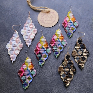 Könnte beinhalten: Eine Sammlung von rautenförmigen Ohrringen mit Perlen in verschiedenen Farben. Die Ohrringe weisen komplizierte Muster in Rosa, Weiß, Gold, Schwarz und mehrfarbigen Perlen auf. Ein Anhänger mit dem Text "Border with Beads" ist ebenfalls sichtbar.