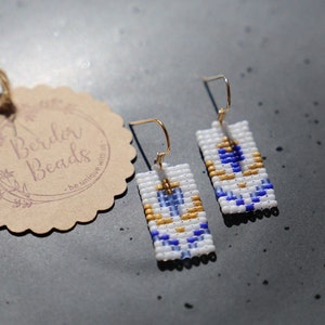 Könnte beinhalten: Ein Paar rechteckige Ohrringe aus weißen, blauen und goldenen Perlen mit goldenen Haken. Die Ohrringe haben ein geometrisches Muster.