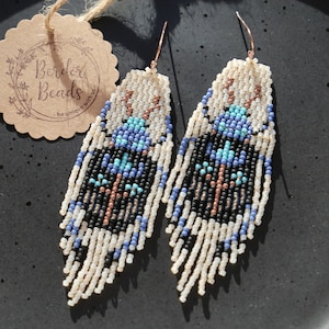 Könnte beinhalten: Ein Paar lange, geperlte Ohrringe mit einem geometrischen Muster in Blautönen, Schwarz, Weiß und Kupfer. Die Ohrringe hängen an einem schwarzen Teller.