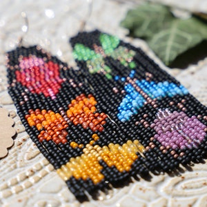 Może przedstawiać: Para długich, koralikowych kolczyków z czarnym tłem i kolorowymi wzorami motyli. Kolczyki są wykonane z małych, okrągłych koralików w odcieniach różowego, pomarańczowego, żółtego, zielonego, niebieskiego i fioletowego.