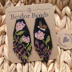 Puede incluir: Un par de pendientes de cuentas negras con diseños florales rosas y verdes. Los pendientes están hechos a mano y presentan un pequeño amuleto de pájaro en un lado. Los pendientes están colgados de una tarjeta marrón con el texto "Bedor Beads" y una etiqueta hecha a mano.