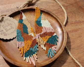 May include: A pair of long, beaded earrings with a geometric pattern in shades of teal, brown, pink, white, and yellow. The earrings are hanging on a wooden plate.