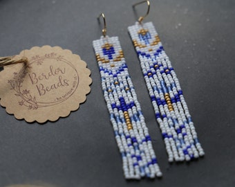 Op de afbeelding: Een paar lange, geperde oorbellen met een geometrisch patroon in wit, blauw en goud. De oorbellen hangen aan gouden haakjes.
