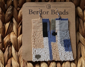 Op de afbeelding: Een paar kralen oorbellen met een geometrisch patroon in tinten blauw, wit, beige en zwart. De oorbellen hangen aan een bruine kaart met de tekst "Berdor Beads".