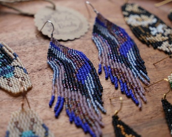 May include: A close-up of several pairs of beaded earrings. The earrings are made with small, colorful beads in shades of blue, black, white, and brown. The earrings are hanging from gold hooks.