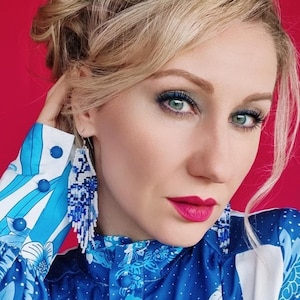 Könnte beinhalten: Eine Frau trägt blau-weiße Perlenohrringe und eine blau gemusterte Bluse. Die Ohrringe sind eine Mischung aus weißen und blauen Perlen. Die Bluse hat ein Blumen- und Federmuster. Der Hintergrund ist einfarbig rot.