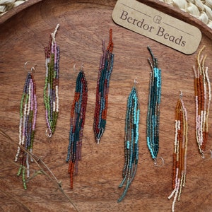 Könnte beinhalten: Eine Sammlung von Perlen-Ohrhängern in verschiedenen Farben, darunter Grün, Lila, Türkis und Braun. Die Ohrringe sind auf einem Holztablett mit einem gewebten Platzset ausgestellt. Der Markenname "Berdor Beads" ist sichtbar.