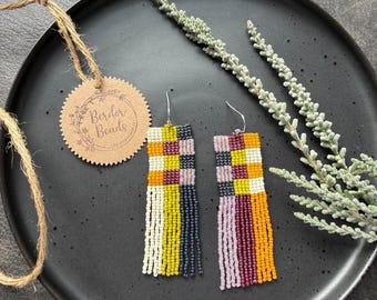 May include: A pair of rectangular beaded earrings featuring a checkered pattern in shades of mustard yellow, dark gray, cream, and mauve. The earrings have long, dangling fringe strands in matching colors and silver-toned metal hooks for attachment.