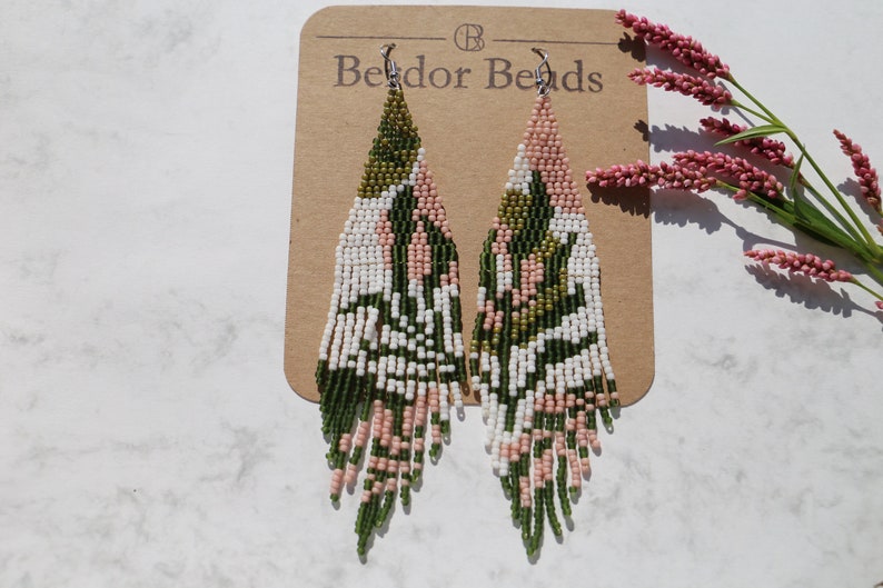 These handwoven beaded fringe earrings feature dark green leaf designs on a white and pink background, offering a trendy, nature-inspired look. A perfect colorful gift for her.