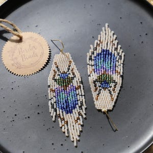Könnte beinhalten: Ein Paar lange, weiße Perlenohrringe mit einem blauen, grünen und violetten Käferdesign. Die Ohrringe hängen an einer schwarzen Platte mit einem braunen Etikett, auf dem "Border Beads" steht.