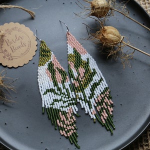 May include: A pair of long, beaded earrings with a green, white, and pink geometric pattern. The earrings are hanging from a black plate.