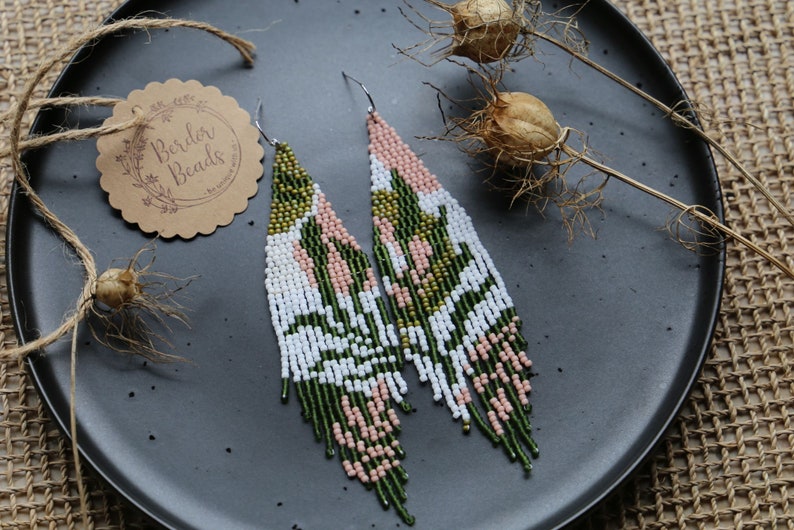 May include: A pair of long, beaded earrings with a green, white, and pink geometric pattern. The earrings are hanging from a black plate.