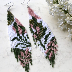 May include: A pair of long, beaded earrings with a geometric pattern in shades of green, white, and pink. The earrings are made with small, seed beads and have a fringe-like design.