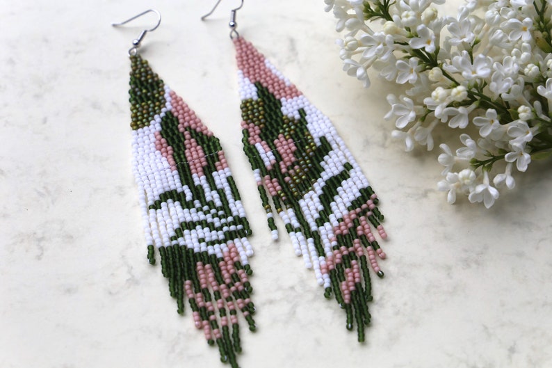 May include: A pair of long, beaded earrings with a geometric pattern in shades of green, white, and pink. The earrings are made with small, seed beads and have a fringe-like design.