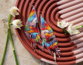 May include: A pair of long, colorful beaded earrings with a geometric pattern. The earrings are hanging on a brown and pink striped surface.