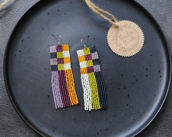 May include: Two beaded earrings with a rectangular design, featuring a mix of colors including purple, yellow, orange, white, and black. The earrings are displayed on a dark gray plate with a tag that reads "Border Beads".