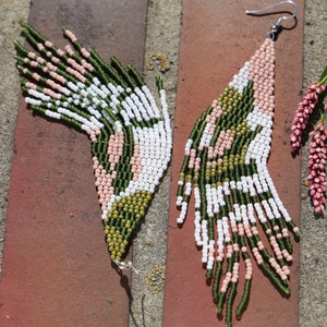 May include: A pair of long, green, white, and pink beaded earrings with a geometric pattern. The earrings are hanging from a brick surface.
