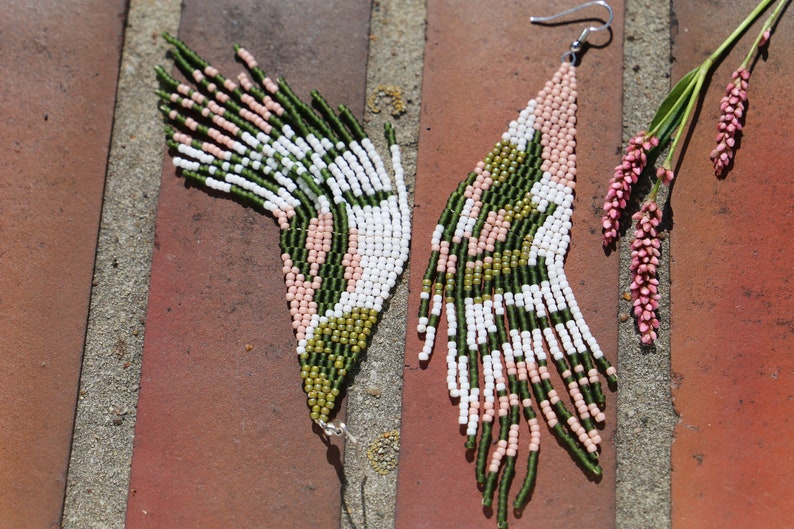 May include: A pair of long, green, white, and pink beaded earrings with a geometric pattern. The earrings are hanging from a brick surface.