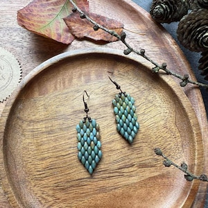 May include: A pair of beaded earrings in shades of teal and green, arranged in a leaf-like pattern. The earrings are displayed on a wooden tray with a tag that reads "Berder Beads". The background includes fall foliage and pine cones.