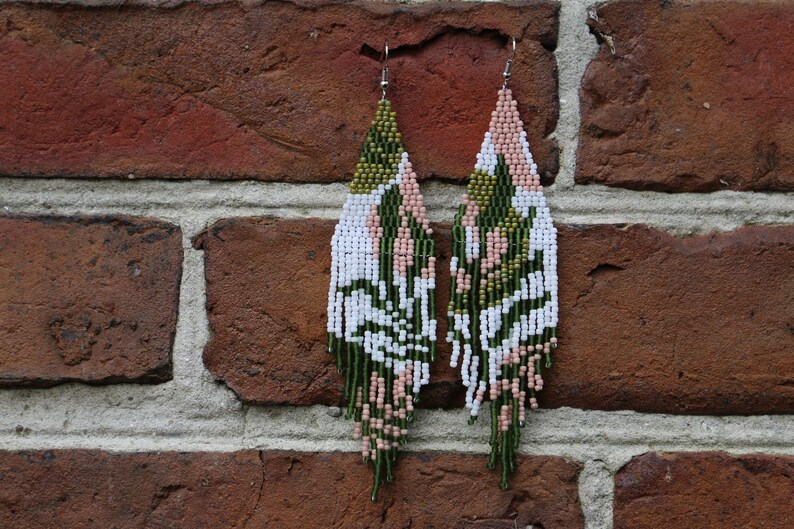 May include: A pair of long, beaded earrings with a geometric pattern in shades of green, white, and pink. The earrings are hanging from a brick wall.