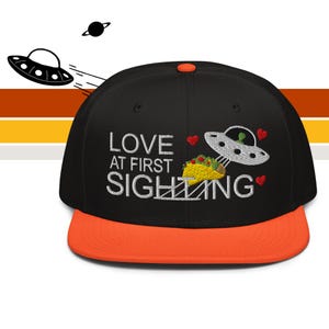 May include: Black baseball cap with an orange peak. The cap has embroidered text reading "LOVE AT FIRST SIGHTING" with a UFO and taco graphic. A small planet and another UFO are in the background.