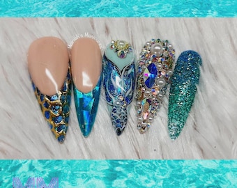 May include: Close-up of five long, pointed, artificial fingernails with different designs. The nails are painted in shades of blue and green with glitter and rhinestones. One nail features a mermaid tail design. The text "Mariah's Manicures" is visible in the image.
