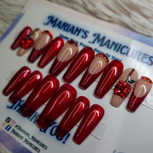 May include: Red glitter press-on nails with a half-moon design. Some nails have a red apple design with rhinestones. The nails are displayed on a white card with the text "MARIAH'S MANICURES".