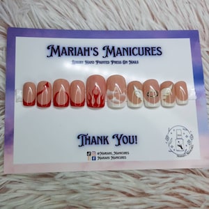 May include: Set of ten press-on nails with a white base and different designs. The designs include red flames, red devil horns, and white angel wings with a gold halo.