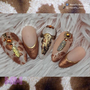 May include: Five nude-colored almond-shaped nails with a brown and white cow print design. The nails are decorated with gold accents, including feathers, a dreamcatcher, and a bull skull. The text "Mariah's Manicures" is written on the nails.