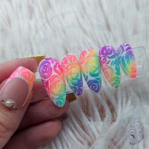 May include: Rainbow-colored artificial nails with a rose design. The nails are painted in a gradient of colors, including pink, orange, yellow, green, and purple. The nails are held in a clear plastic holder.