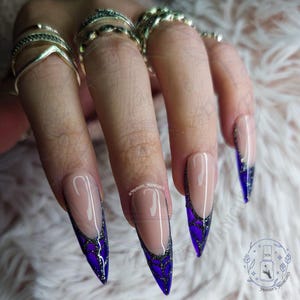 May include: Long, pointed fingernails with a French manicure design. The nails feature a nude base with a white outline and a deep purple and black glitter design at the tips. The image includes the text "Marian's Manicures".