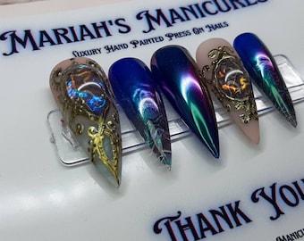 May include: Five press-on nails with a variety of designs. The nails are in a stiletto shape and have a variety of colors, including blue, green, and iridescent. The nails are decorated with gold accents and a design that looks like a mirror. The text "MARIAH'S MANICURES" and "LUXURY HAND PAINTED PRESS ON NAILS" is visible at the top of the image. The text "THANK YOU" is visible at the bottom of the image.