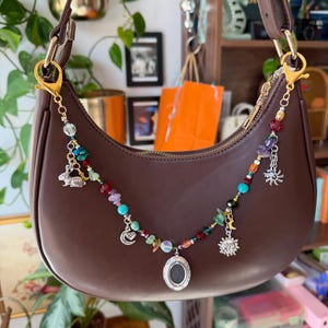 May include: A dark brown crescent-shaped shoulder bag featuring a decorative beaded chain attached to the gold-tone clasps. The chain includes colourful gemstone beads, a silver rabbit charm, a crescent moon charm, a sun charm, and a central oval locket.