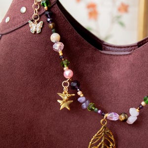May include: A decorative bag charm featuring a gold-tone metal ring, a white butterfly charm, a gold-tone hummingbird and star charm, and a large gold-tone leaf pendant. The chain is composed of various colorful beads, including pink, green, purple, and brown stones.