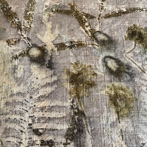 May include: Close-up of a textile with a botanical print. The fabric has a light gray background with olive green and brown leaf and plant designs. The top edge has a fringe.