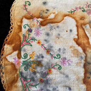 May include: A vintage, off-white handkerchief with embroidered floral designs in pink, yellow, and blue. The edges are trimmed with a crocheted border. The fabric has brown and gray discoloration.