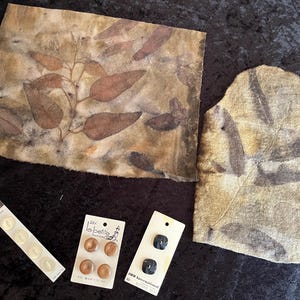 May include: An assortment of craft supplies including fabric pieces with leaf imprints in shades of brown and green, along with three sets of buttons in various styles and colors. The buttons are on white cards.