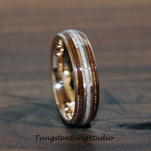 May include: A gold-coloured ring featuring a central band of white opal, framed by bands of dark wood. The ring has a polished finish. The text "TungstenRingStudio" is visible at the bottom.