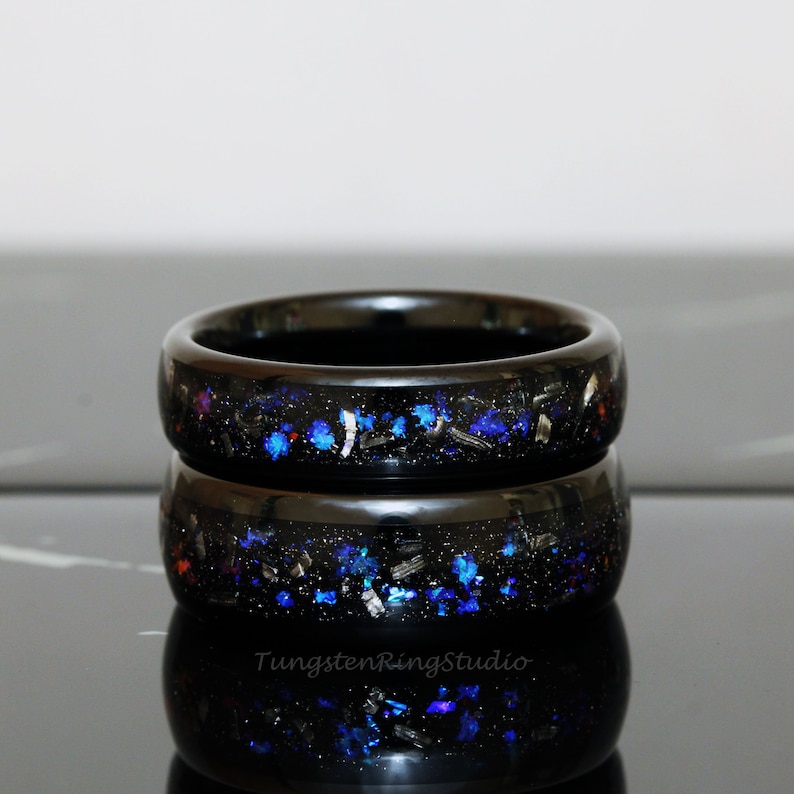 May include: Two black tungsten carbide rings with blue and silver glitter embedded in the black resin. The rings are stacked on top of each other.