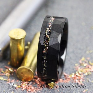 May include: Black tungsten ring with a faceted design and a central inlay of mixed metals. The ring features a dark, textured surface and is accented with gold and copper-colored flecks. The ring is displayed near brass bullet casings.