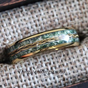 May include: A gold ring featuring a green stone inlay and a braided gold band. The ring is displayed on a textured, neutral-toned surface. The text "TungstenRingstudio" is visible at the bottom of the image.