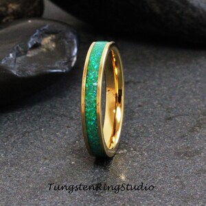 May include: A gold-colored ring with a vibrant green inlay. The ring has a polished finish and a band of gold. The green inlay is made of small, sparkling pieces. The text "TungstenRingStudio" is at the bottom.