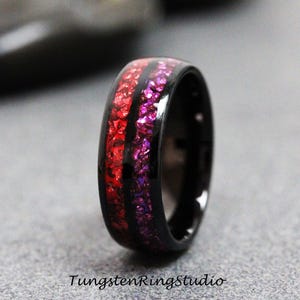 Puede incluir: Un anillo negro con una vibrante incrustación roja y morada. El anillo presenta un acabado brillante y una banda negra. El texto "TungstenRingStudio" está en la parte inferior de la imagen.