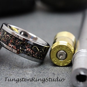 May include: A tungsten ring with a black, copper, and gold inlay, next to a bullet casing and a silver tube. The ring has a polished silver band and the text "TungstenRingStudio" is at the bottom.