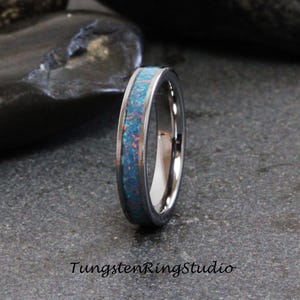 Può includere: Un anello color argento con una fascia di opale blu e rosso. L'anello è posto su una superficie grigio scuro. Il testo "TungstenRingStudio" è in basso nell'immagine.