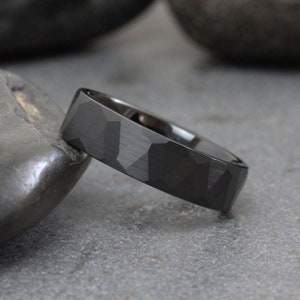 May include: A black wedding band with a geometric patterned design. The band has a matte finish and a slightly rounded edge.